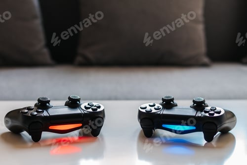 Preview: Photo of two gamepads with selective focus, on white table with gray sofa in the background