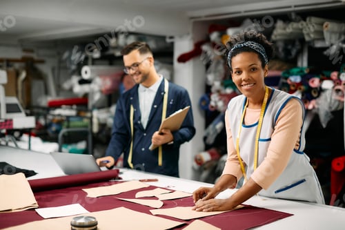 Preview: Happy African American dressmaker working with paper pattern and chalk at tailor's.