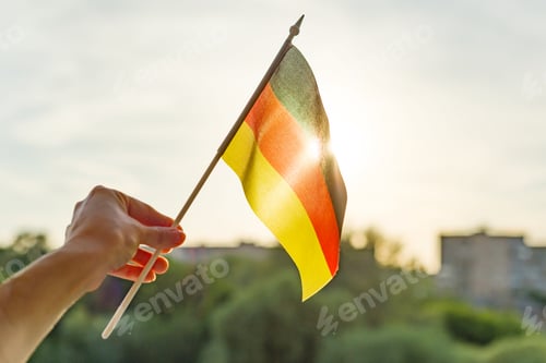 Preview: Hand holding Germany flag an open window. Background blue sky, silhouette of the city, sunset
