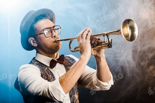 Preview: stylish jazzman in hat and eyeglasses playing on trumpet on stage with dramatic lighting and smoke