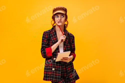 Preview: Thoughtful lady in trendy jacket and cap holds pen and notebook on orange background. Red haired wo