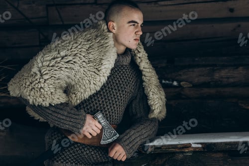 Preview: Ancient viking warrior with steel axe posing near wooden hut in scandinavian forest