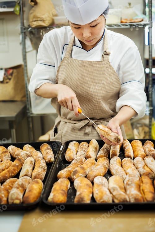 Preview: Woman wearing chef's hat and apron working in a bakery, slicing freshly baked rolls on large trays.