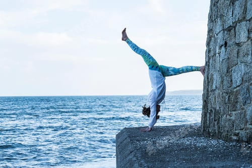 Preview: Young woman doing handstand on a wall by the ocean, one leg raised.