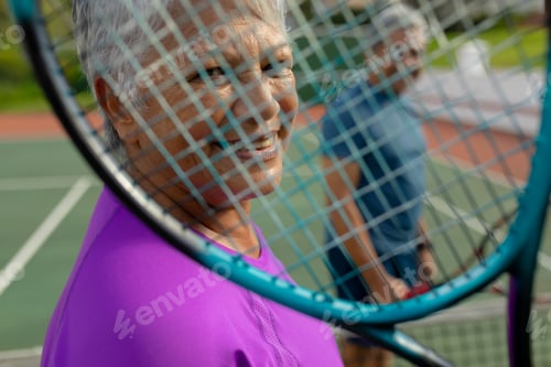 Preview: Close-up portrait of biracial smiling senior woman with man looking through tennis racket in court