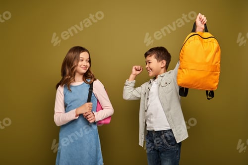 Preview: Excited multiracial boy holding backpack and showing yes gesture near friend in dress standing and