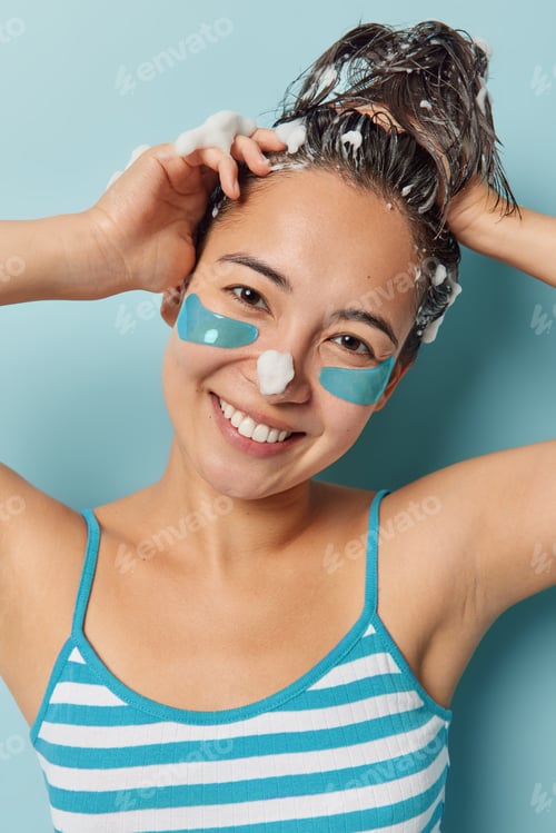 Preview: Cheerful young Asian woman washes hair with shampoo applies hydrogel patches under eyes for delicate