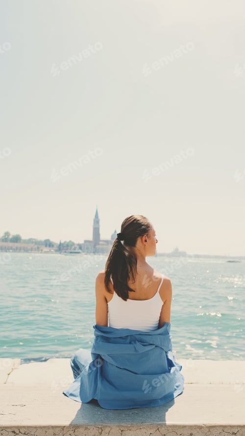 Preview: Beautiful girl with long brown hair, dressed in blue shirt, sits and looks at sea, Back view
