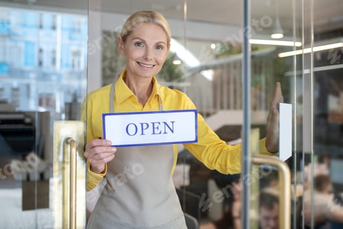 Preview: Blonde woman opening glass door, holding sign saying open