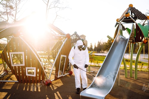 Preview: Man wearing protective suit disinfecting the playground in the sun with spray. Covid -19. Cleaning.