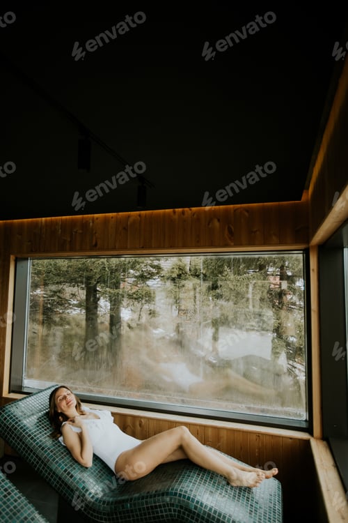 Preview: Woman lying on a warm stone bed near a large window overlooking trees and nature