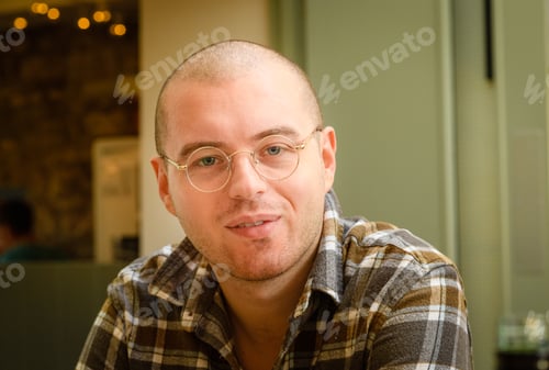 Preview: a young man wearing glasses and a check shirt with a shaved head and beard sitting in a restaurant