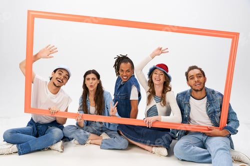 Preview: cheerful and trendy multiethnic friends sitting with picture frame and gesturing on grey backdrop