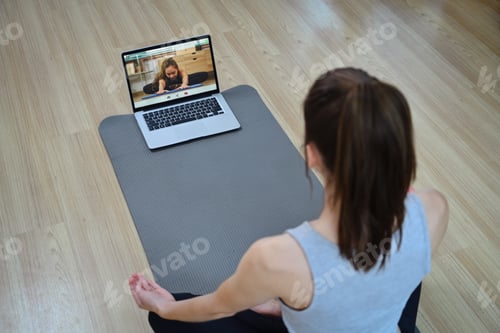 Preview: Back view of young woman sitting on yoga mat watching active sports workout video lesson on laptop