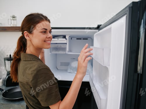 Preview: Woman smiling with teeth looking into camera in kitchen at home opened freezer empty with ice inside