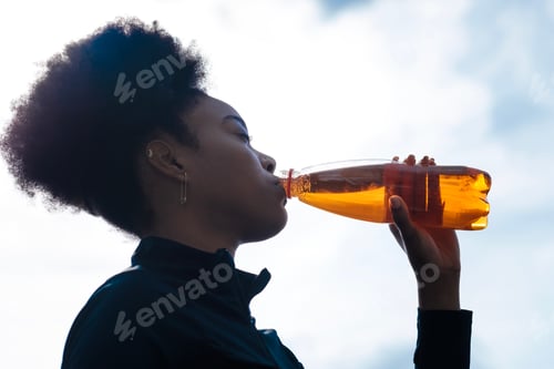 Preview: Silhouette of young fit black woman drinking a bottle of energy drink.