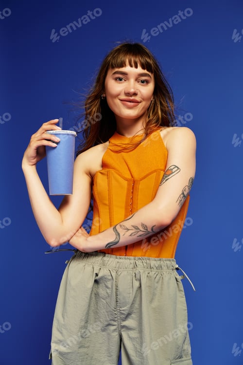 Preview: Stylish young woman holding a trendy drink against a vibrant blue background