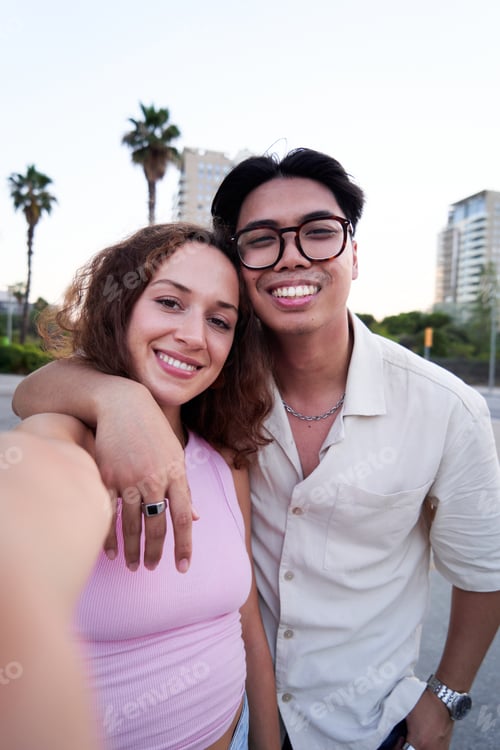 Preview: Multiracial couple using phone to take a selfie looking at camera smiling standing outdoors.