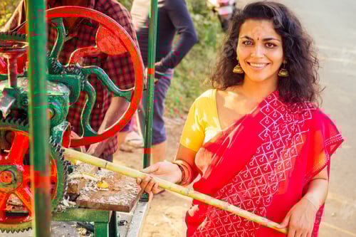 Vista previa: hermosa mujer india con sari rojo de pie cerca de una máquina para hacer jugo de caña de azúcar
