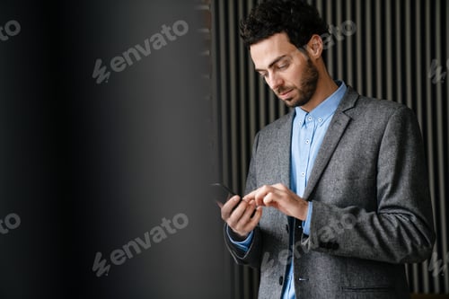 Preview: Confident focused businessman in jacket using mobile phone indoors