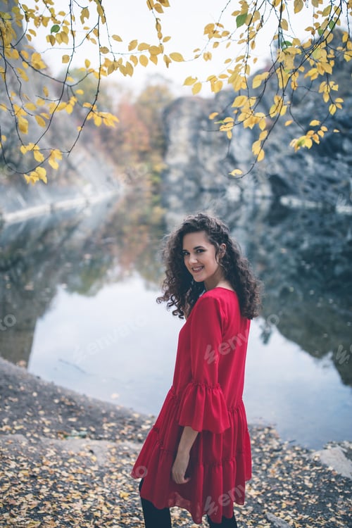 Preview: Beautiful curly hair girl in a red dress standing at the lake and smiling