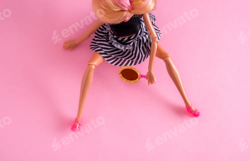 Preview: Top view of plastic doll sitting with her legs spread and holding mirror on pink background.