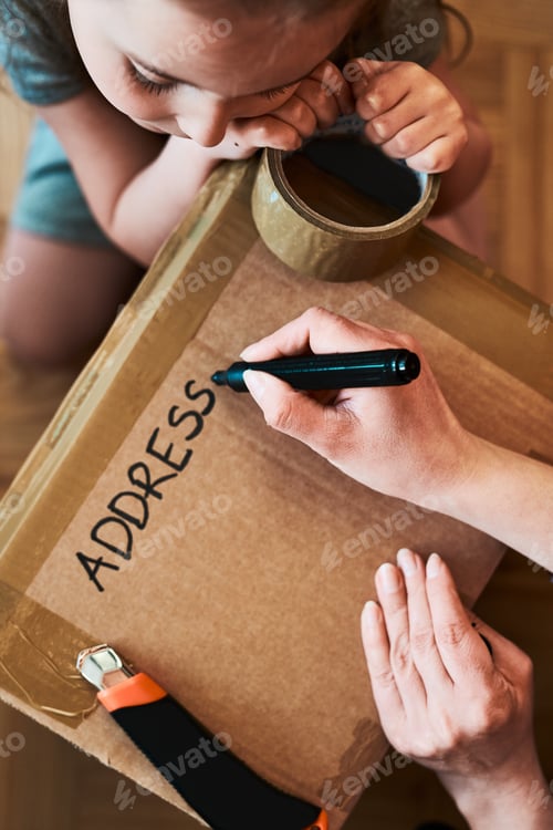 Preview: Woman writing an address on a cardboard box parcel in room at home. Little girl helping her mother