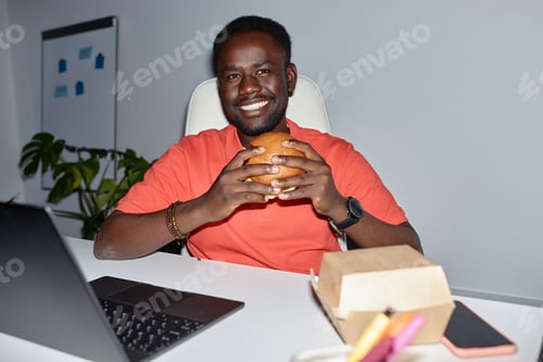 Preview: Smiling black man eating burger at workplace indoors