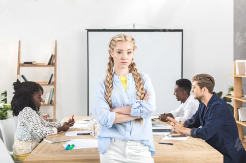 Preview: portrait of upset businesswoman with arms crossed looking at camera and colleagues working behind in