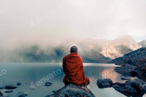 Preview: Tibetan monk from back sitting on the stone near the water in the background of foggy mountains