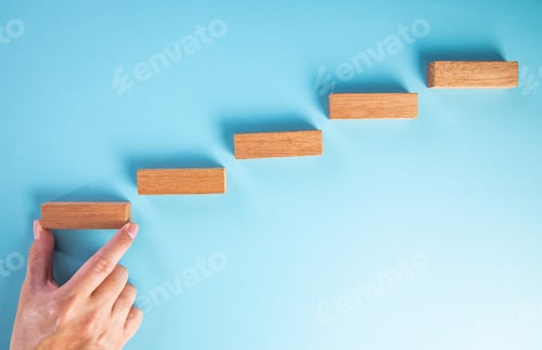 Preview: Hand placing wooden block on ascending steps towards success on blue background