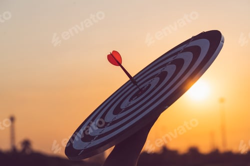 Preview: Close up red dart arrow hitting target center dartboard on sunset background.