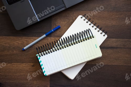 Preview: Top view of a wooden desk with a notebook, and a pen next to a laptop computer.