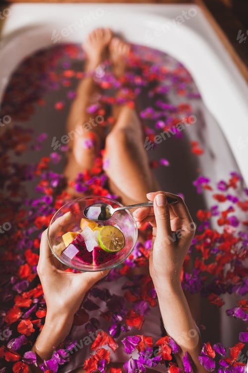 Preview: Woman legs in flower bath in spa. Hands holding plate with fresh fruit salad, view from above