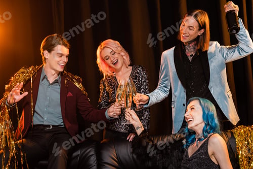 Preview: excited queer friends clinking champagne glasses near golden tinsel on dark background with lighting