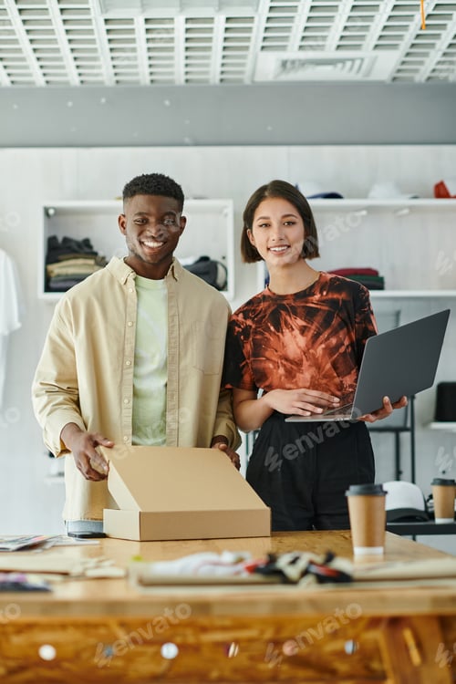 Preview: joyful multicultural entrepreneurs with laptop and carton box looking at camera in print studio
