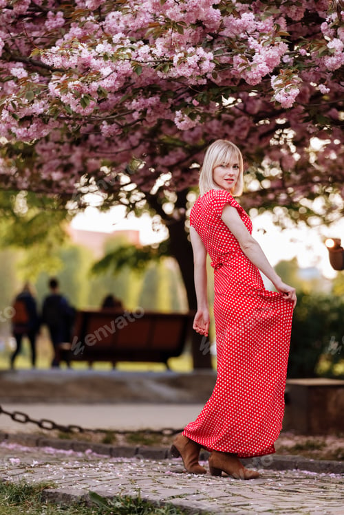 Preview: beautiful woman in red dress in the city near flowering trees. Outdoor portrait of young beautiful