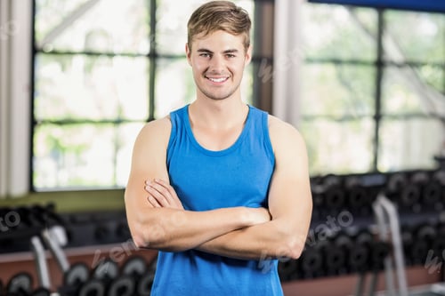Preview: Man standing with arms crossed in gym area wearing blue sleeveless top with dumbbells and windows