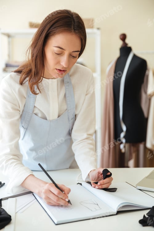 Preview: Cocentrated woman seamstress in blue apron drawing new scetch