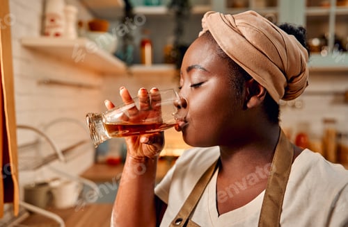 Preview: Household chores. Side view of black curvy woman in traditional turban enjoying refreshing drink wit