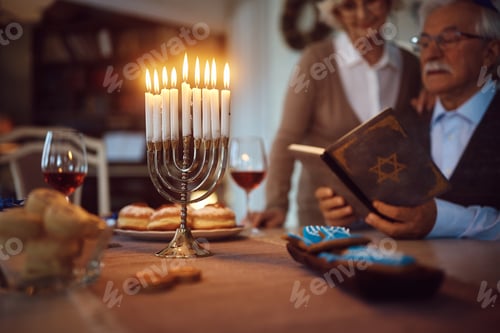 Preview: Burning candles in menorah with senior couple reading Tanakh during Hanukkah celebration.