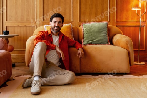 Preview: Smiling bearded man sitting on floor in cozy home interior, fashion model posing