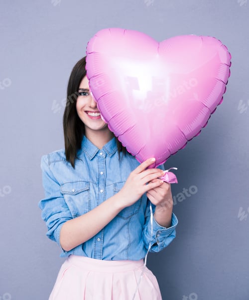 Preview: Smiling Woman Covering Her Eye With Heart Shaped Balloon Over Gray Background. Looking At Camera