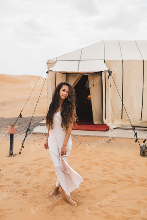 Preview: Portrait of beautiful oriental young brunette woman in white dress. Glamping tent in moroccan desert