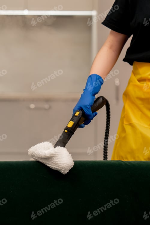 Preview: apartment cleaner in yellow apron does a steam cleaning of a green sofa with professional equipment