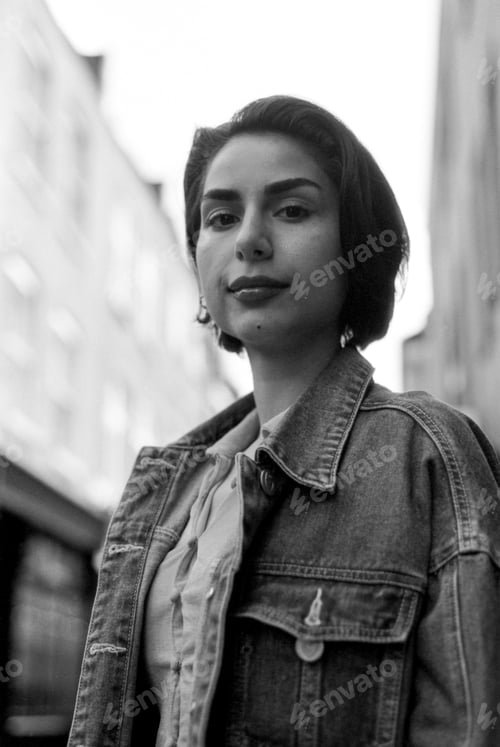 Preview: Young woman wearing denim jacket in urban setting, grainy monochrome portrait