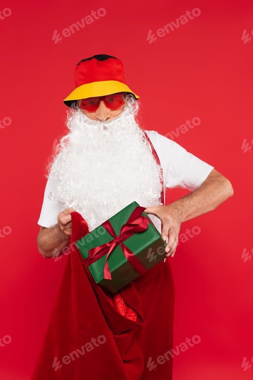 Preview: Santa claus in panama and sunglasses putting present with bow in sack isolated on red