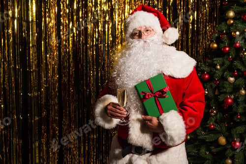 Preview: Santa claus holding present and champagne near christmas tree and tinsel