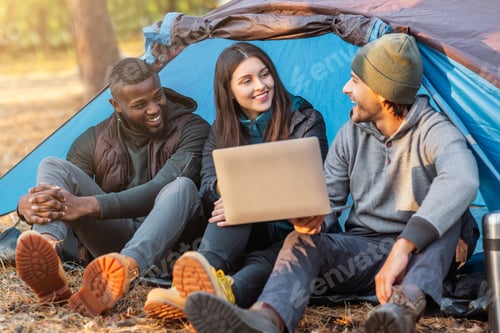 Preview: Three happy friends sitting in camping tent and using laptop