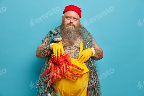Preview: Dejected bearded stout man mariner being in bad mood, holds red octopus and wears overalls. Offended